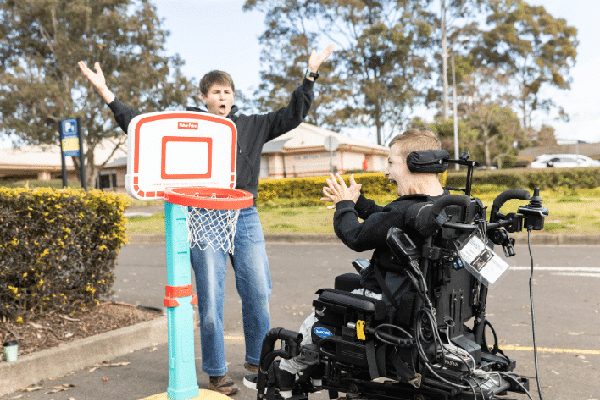 A boy in a power wheelchair and his therapist celebrating the achievement of a goal, to shoot a basketball into the hoop.