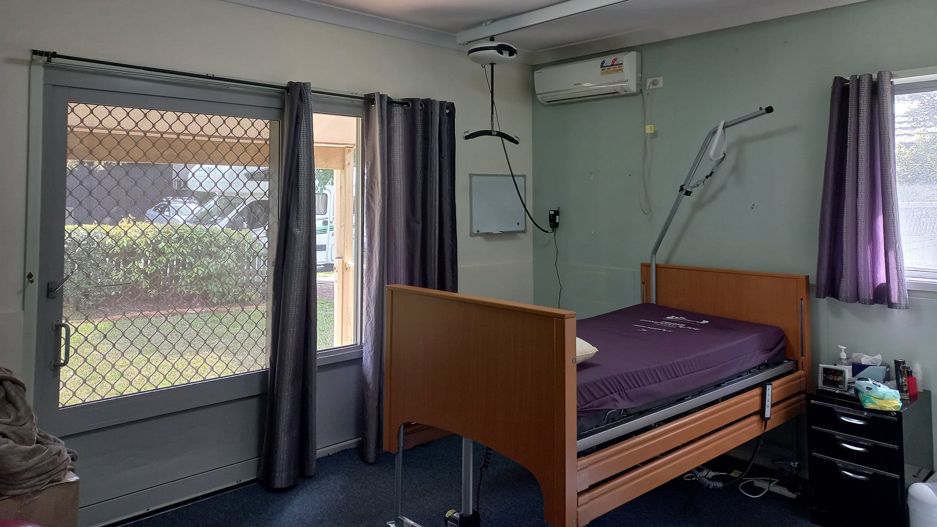 A bedroom with a large secure window, a bed with hoist supports and air conditioning.
