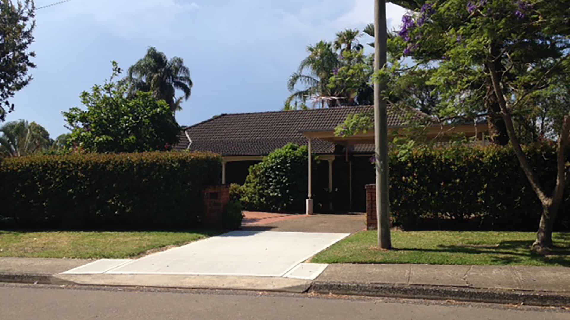 Street view of the Terrey Hills 1 property, a tall hedge sits in front of the house, the driveway leads to a covered car spot that connects to the house