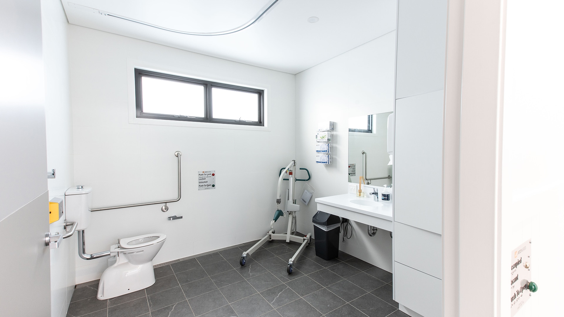 A bathroom with toilet underneath two windows, mobility devices and a sink area.