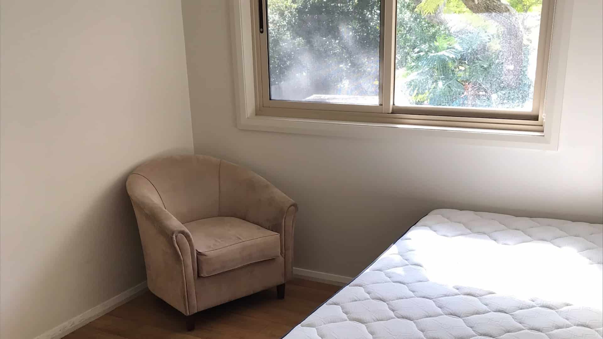 View of a bedroom with a bed and chair below a window.