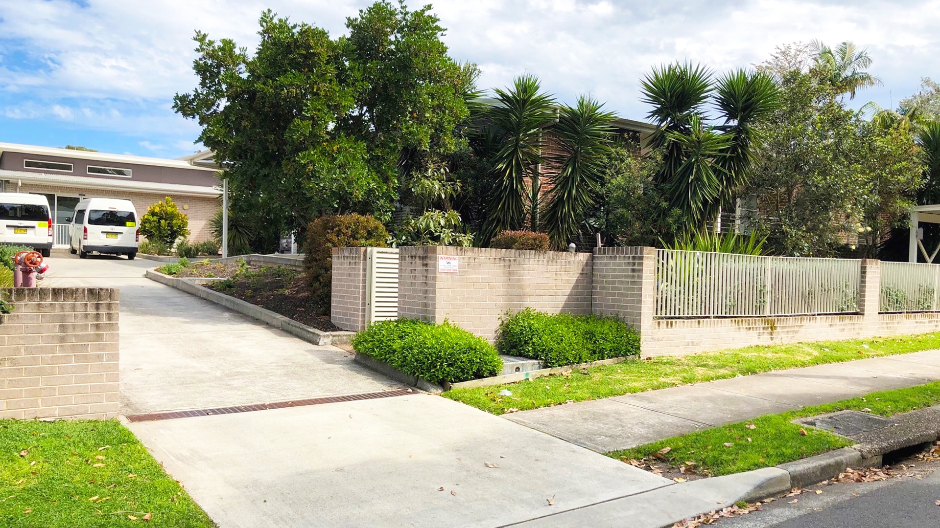 Front view of the Narraweena 2 property. Brick wall with green leafy plants behind it and a driveway leading up to parked vans with a building behind it.