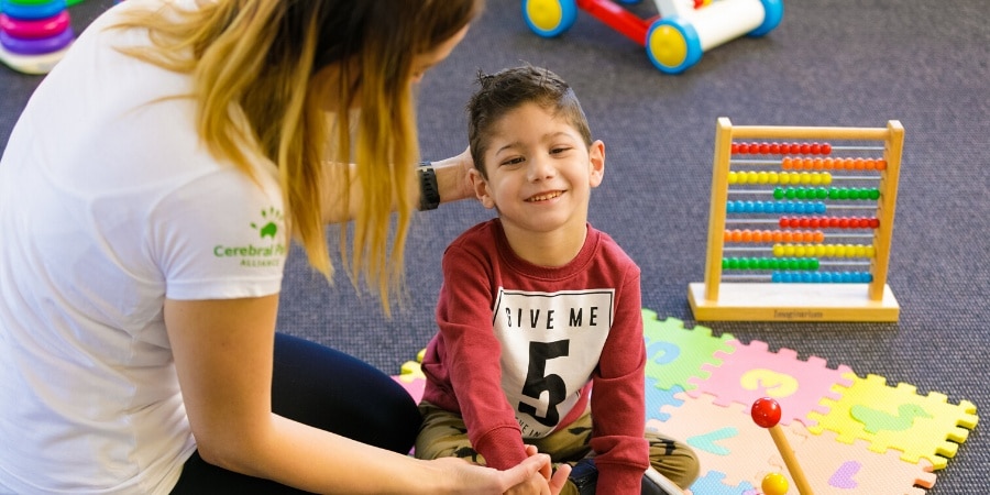 A young boy sitting on a playmat surrounded by toys with a therapy worker supporting him