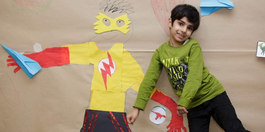 a young boy smiling, standing in front of a paper artwork of a superhero