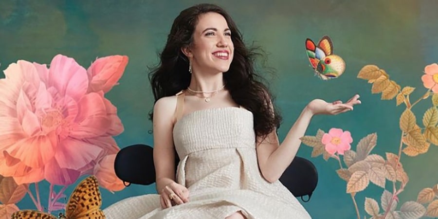 White woman with long black hair sitting down smiling with a butterfly and flowers in the background.