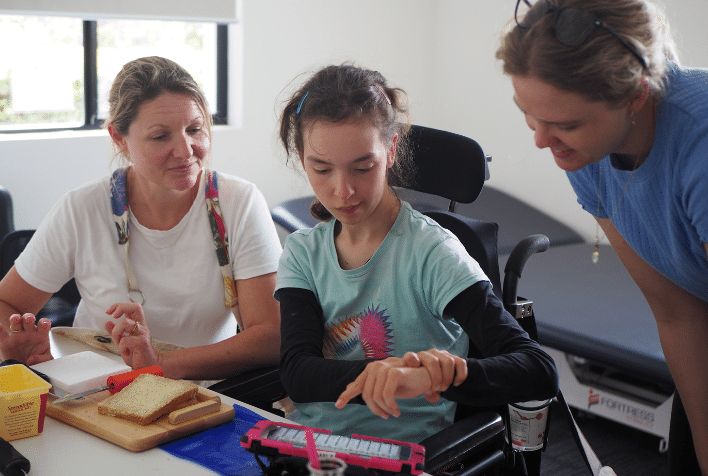 A young female sitting in a wheelchair using a communication device while making a sandwich with friends.