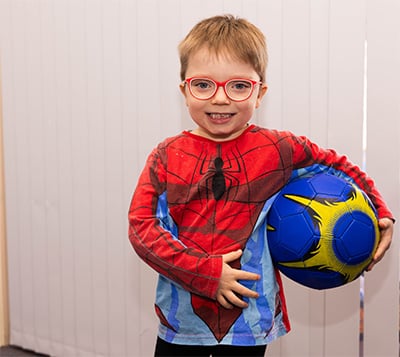 Happy boy in a spiderman suit and red glasses holding a soccer ball.