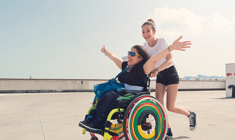 A girl in a wheelchair smiling with her arms outstretched with a friend behind her pushing the wheelchair and smiling