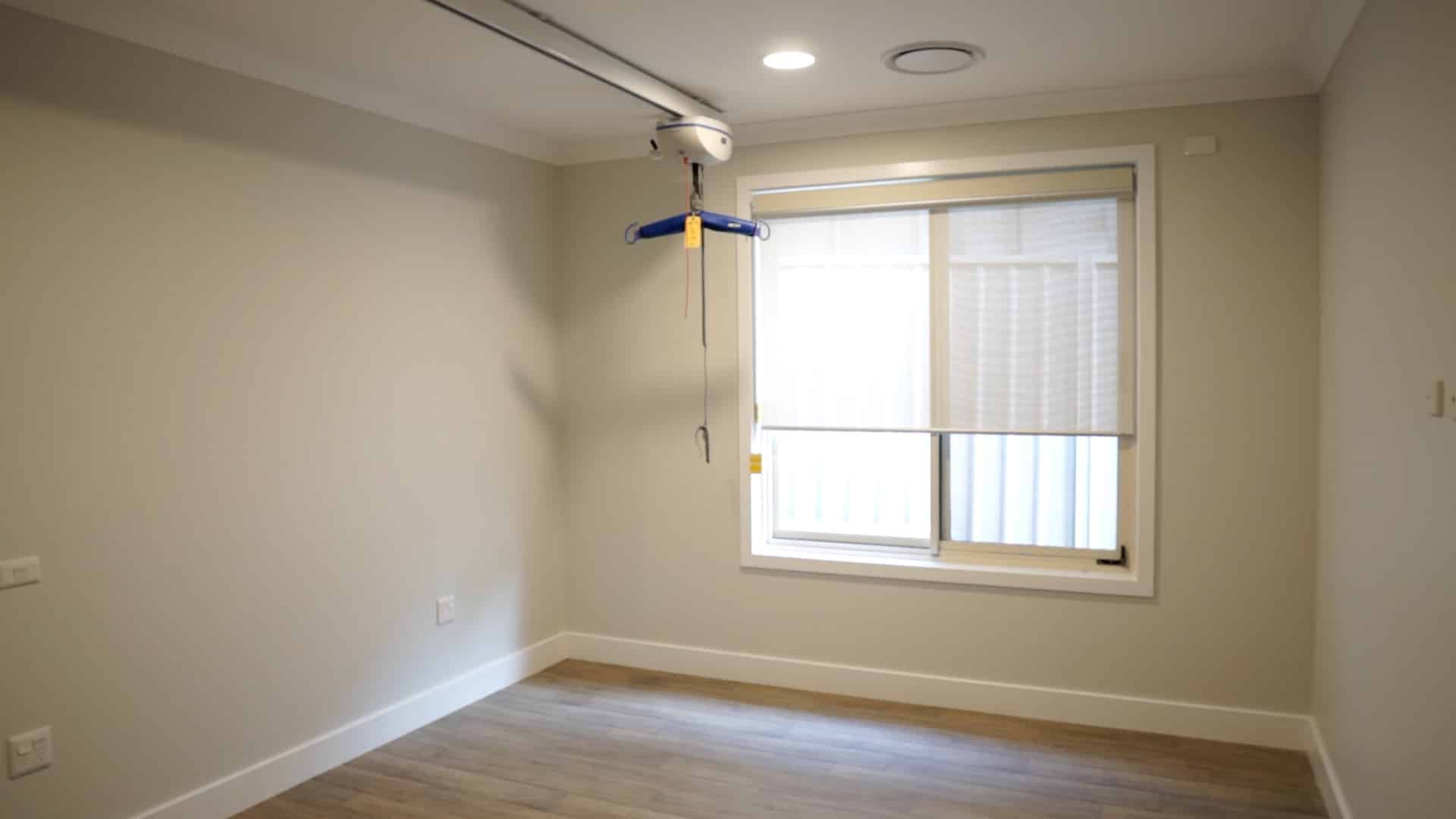 View of a bedroom with a window, floor boards and a ceiling hoist.