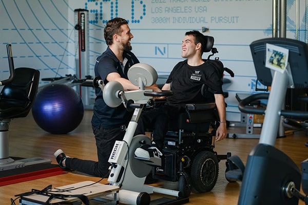 A male CPA client sitting in a wheelchair pedaling a bike machine with his physio
