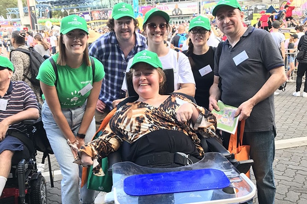 A group of volunteers at an event standing with a lady sitting in a wheelchair. All people are wearing hats