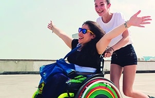 A girl in a wheelchair smiling with her arms outstretched with a friend behind her pushing the wheelchair and smiling