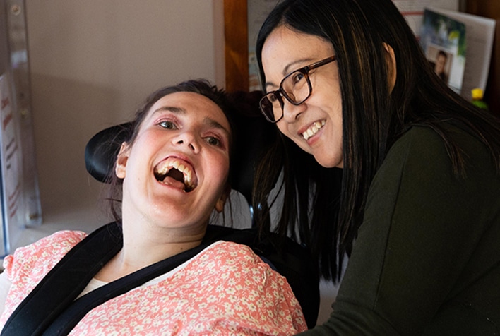 A lady sitting in a wheelchair next to her support worker, both smiling