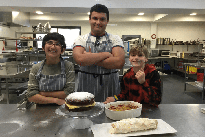 Three young males standing together in an industrial kitchen with cakes they have made in front of them