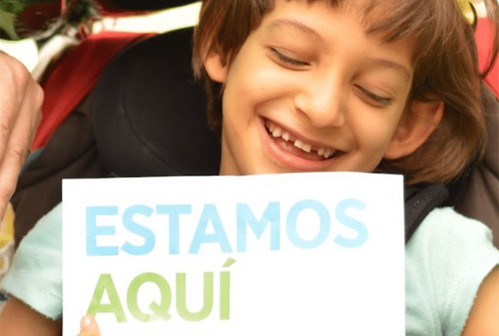 Young boy smiling with a sign that says Estamos Aqui