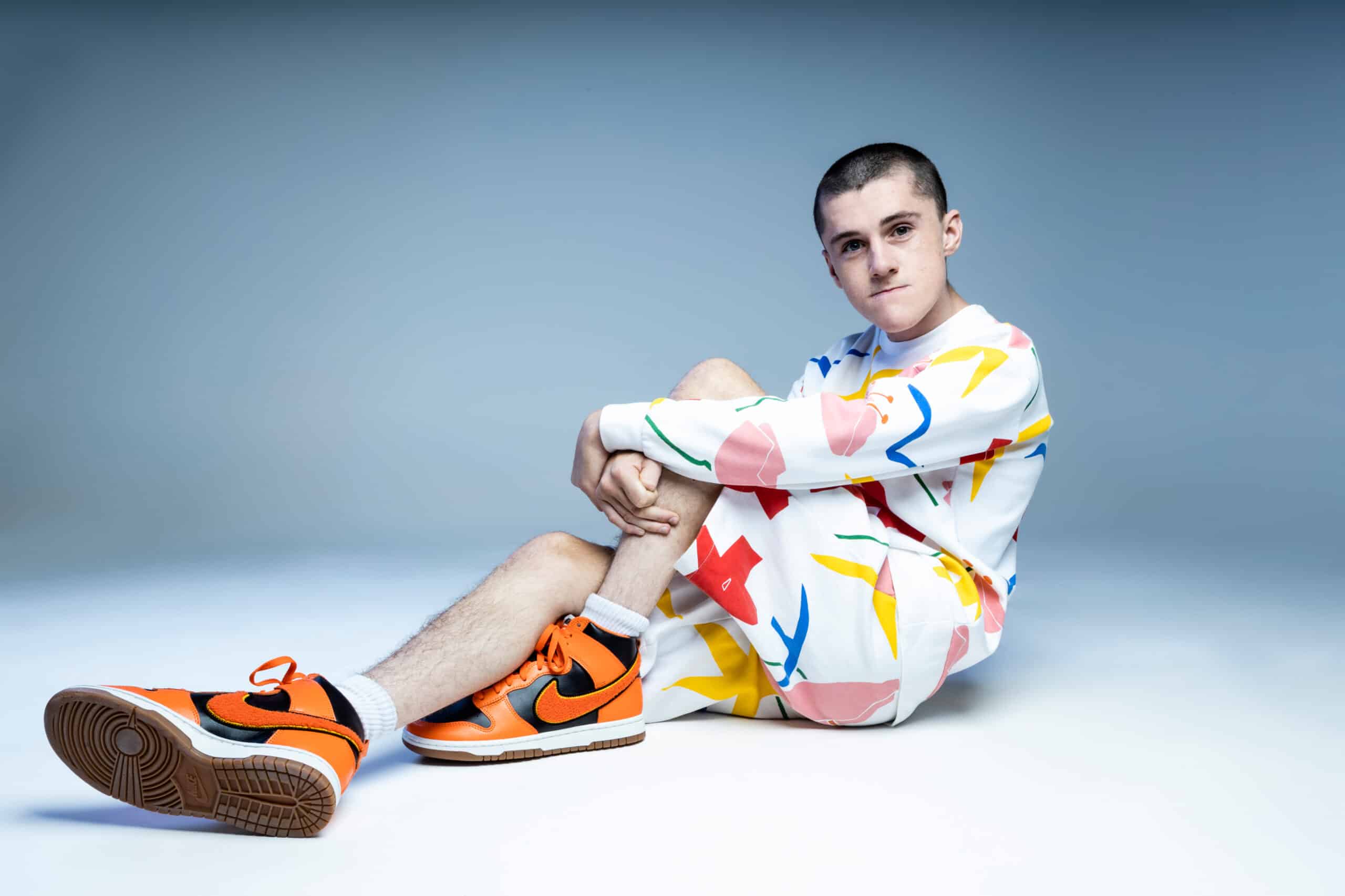 DJ Cooper sitting on the floor with arms folded over legs wearing a bright outfit.