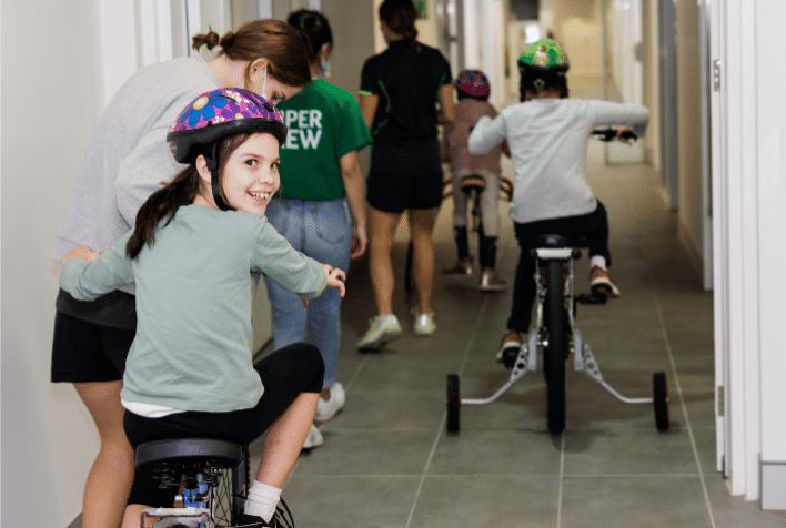 three children riding bikes down a hallway with support workers walking alongside them
