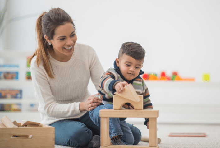 A mother sitting in a playroom with her toddler son on her lap building with blocks