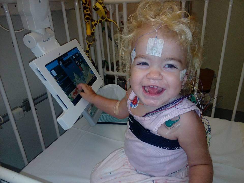 A baby in a hospital bed with medical sensors taped to her chest and face, smiling and playing on a computer device.