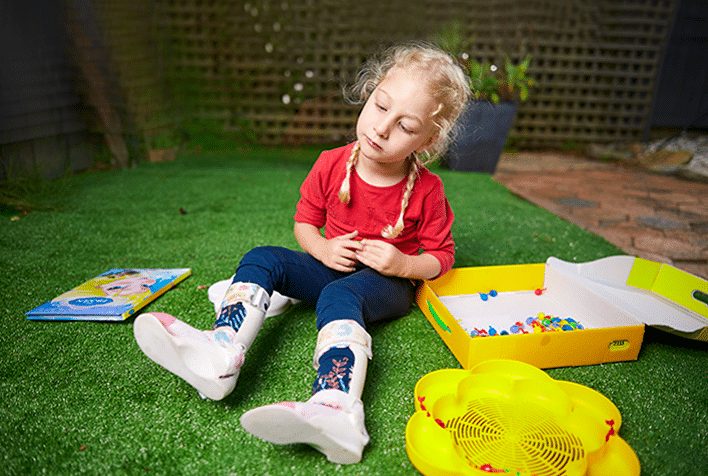 Little girl with blonde pigtails sitting on the ground with her legs outstretched wearing leg brace devices on her lower legs, surrounded by toys
