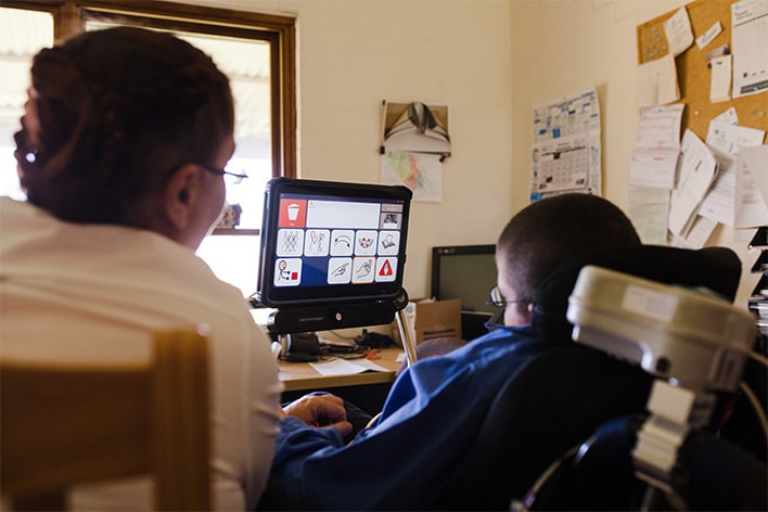 A young child sitting in a wheelchair using eye-gaze technology on a screen, next to an adult