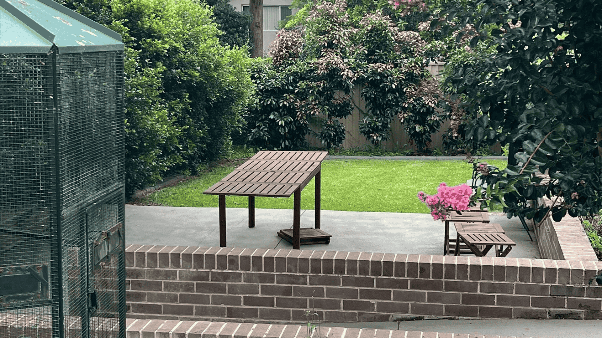 View of the backyard, showing a paved area with dining table and chairs, in front of a grassed area surrounded by trees.