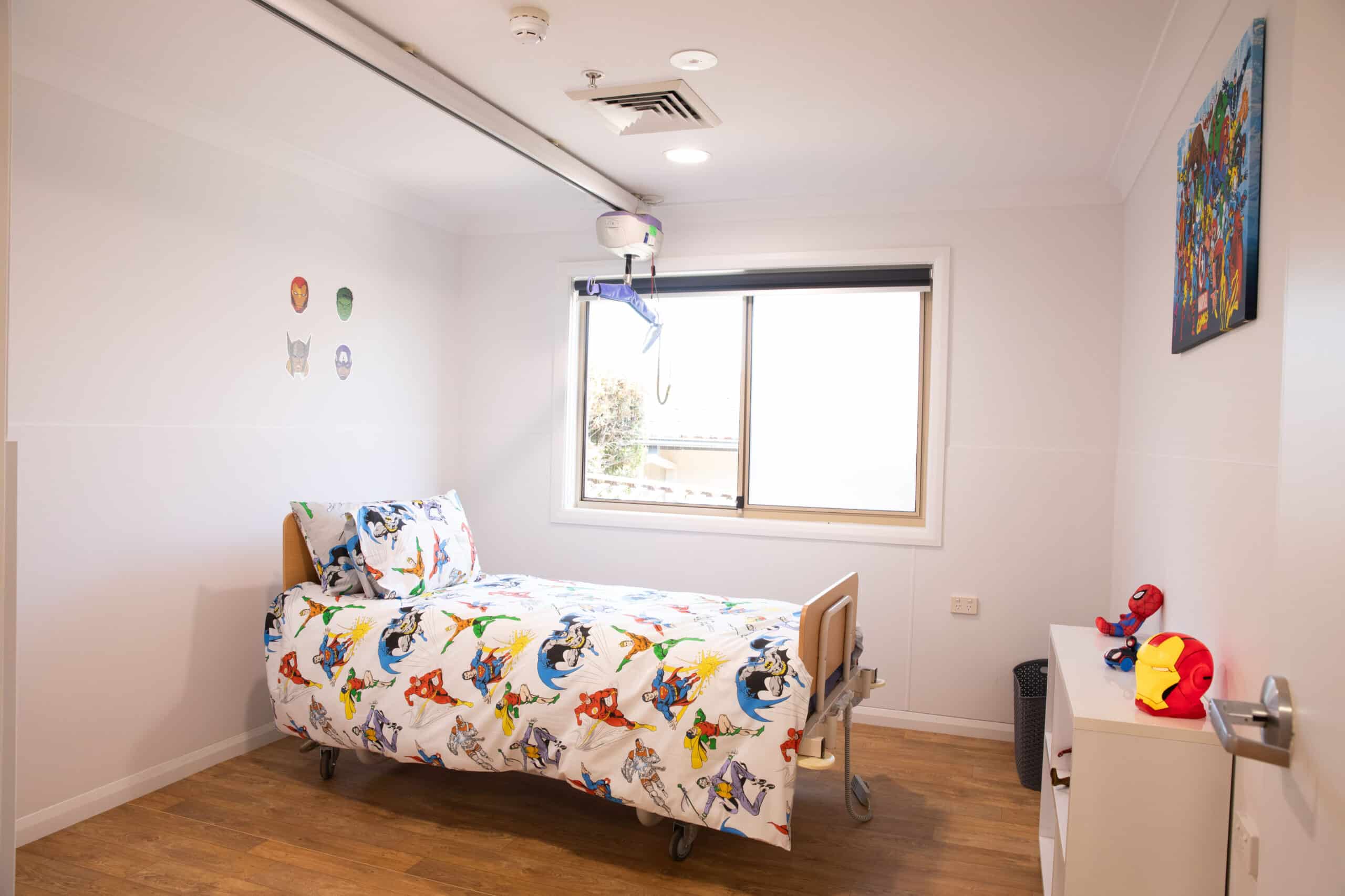 A child's bedroom with a single bed in the middle of the room, a window and some shelves with superhero toys on it.