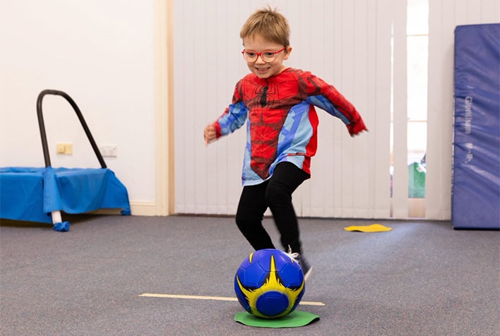 little boy wearing glasses and a spiderman top kicking a soccer ball