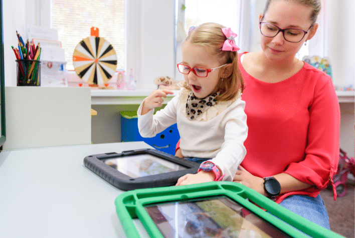 Little girl wearing glasses looking down at an ipad on a desk while sitting on her mum's lap