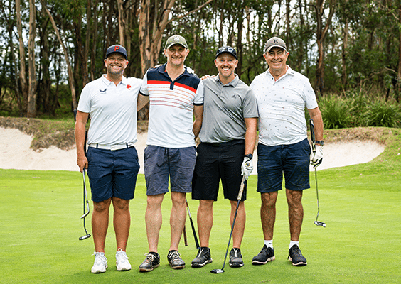 Four white middle aged males standing next to each other on a golf course, dressed in golf attire and holding golf clubs