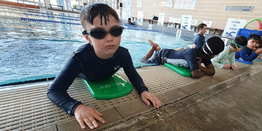 Boy at swimming pool wearing a black swim top and goggles with other kids in the background