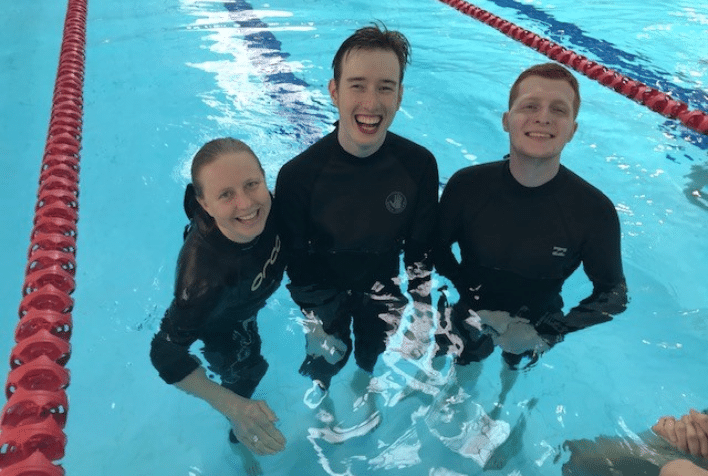 A group of 3 young adults, smiling standing in a pool wearing wetsuits