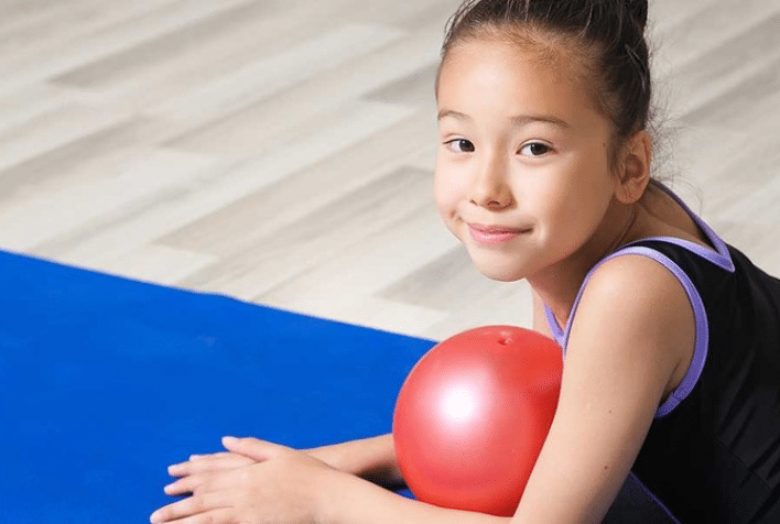 A young girl dressed for a sports class smiling holding a ball.