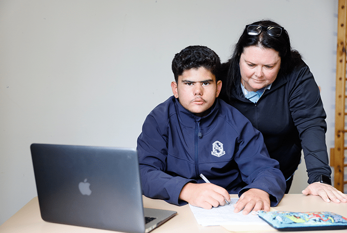 A teenage boy sitting at a table doing school work while his mother looks over his shoulder at what he is doing.