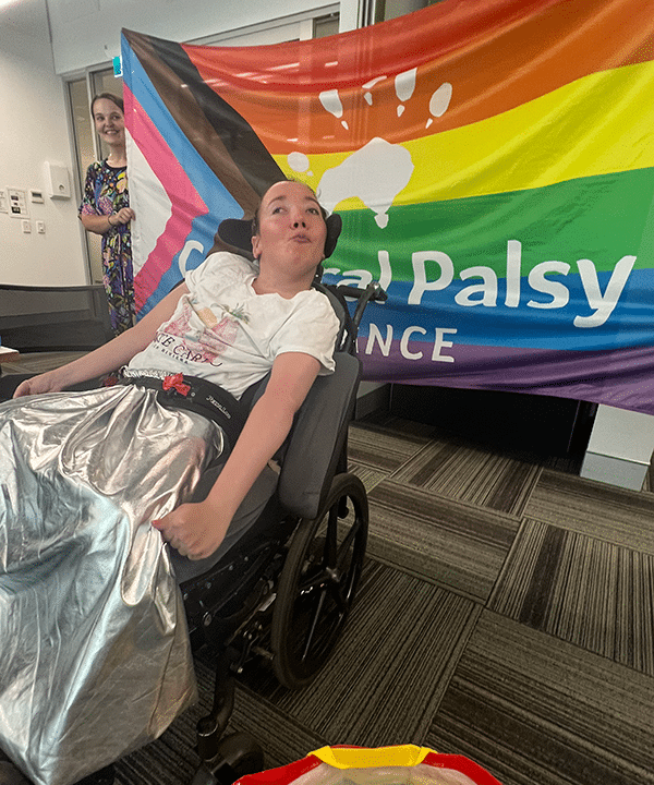 A person in a wheelchair posing in front of the CPA rainbow mardi gras flag
