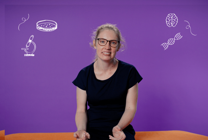 White woman wearing glasses and a black top with blonde hair sitting on an orange seat, on a purple background.