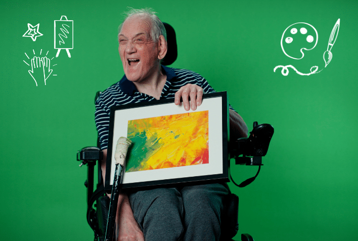 White elderly man holding painting sitting in a wheelchair laughing