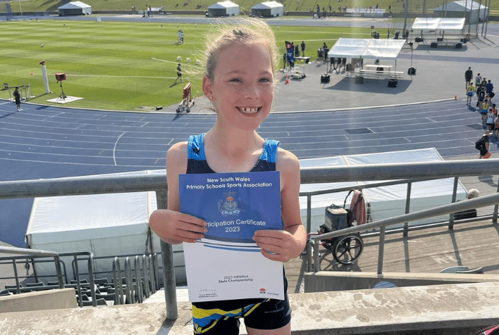 A young girl smiling holding a certificate in front of a running track