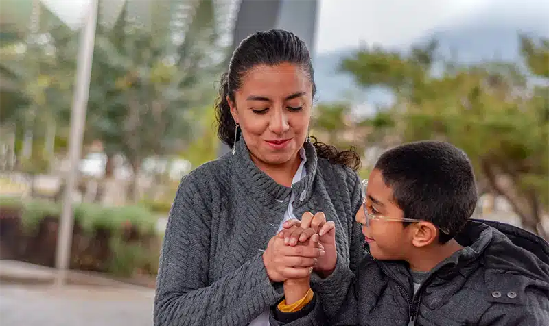 A tender moment as a boy with cerebral palsy holds his mother's hand during an outdoor interaction.