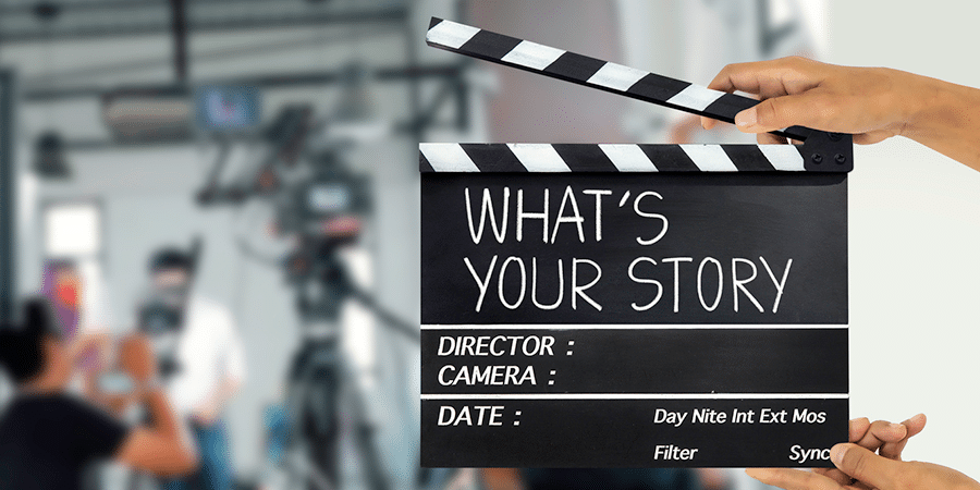 A clapperboard held up with the words 'What's your story' printed on it. A blurred view of a film set is in the background.