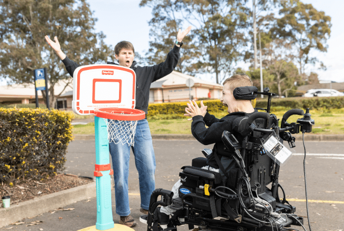 A boy in a power wheelchair and his therapist celebrating the achievement of a goal, to shoot a basketball into the hoop.