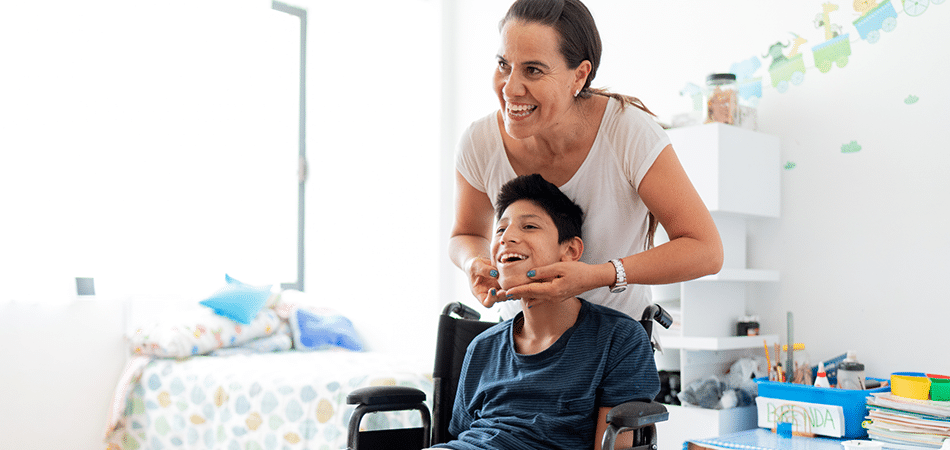 A young boy sitting in a wheelchair with his mother standing behind him with his hands on his face, both smiling towards a mirror.