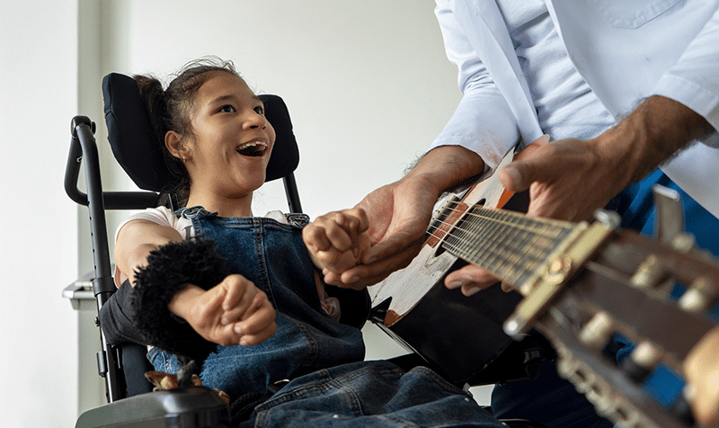 A teenage girl in a wheen chair learning to play guitar.