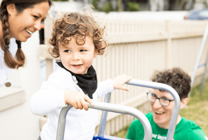 A toddler standing at the top of a slippery-dip with a male and female therapist looking on.