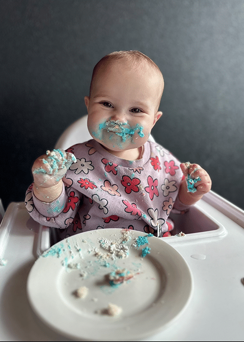 Baby Sadie sitting in a highchair, smiling enjoying cake.