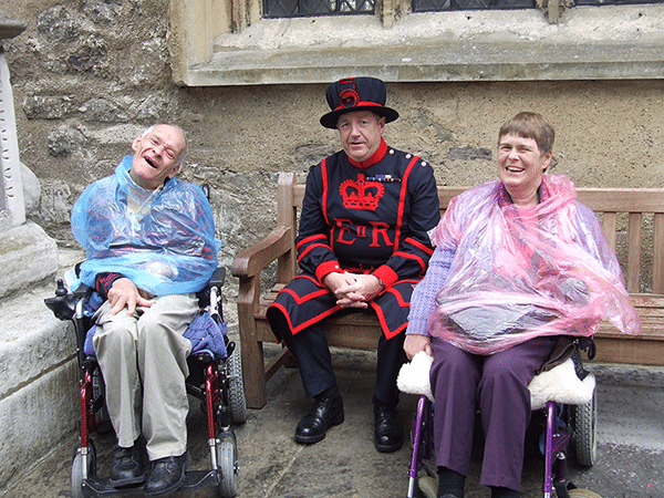 Lindsay and Maria sitting in their wheelchairs wearing rain ponchos, posing with a guard at the Tower of London