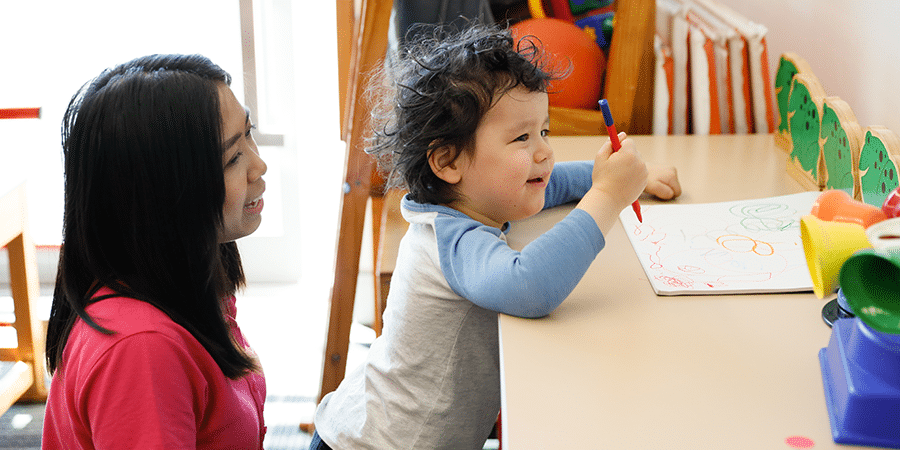 A young boy standing at a table holding a pencil with a therapist sitting behind him watching on