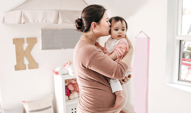 A pregnant mother stands in a nursery holding her baby girl and kissing her on the cheek
