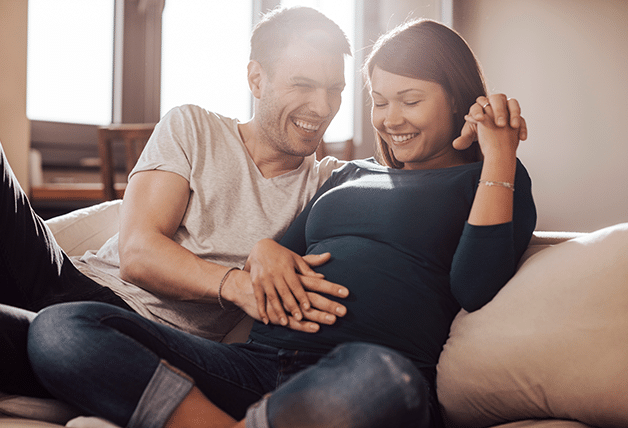 A pregnant woman sits on a couch in the sunlight with her husband next to her, both smiling and placing a hand on her stomach.