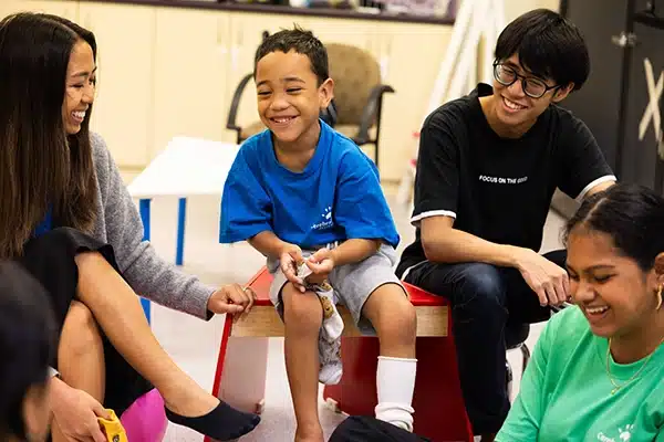 A young boy sitting on a bench holding a sock he is about to put on, smiling to the group of adults gathered around him.